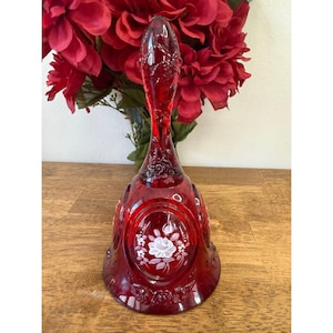 Puede incluir: Una campana de cristal rojo rubí con un mango largo y detalles florales. La campana presenta un diseño ovalado con una rosa blanca y rosa. La campana es un objeto decorativo.