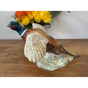 May include: A ceramic pheasant ornament with detailed plumage in shades of brown, cream, and green. The bird has a blue-green head and a red eye patch. The ornament is perched on a base with green foliage.