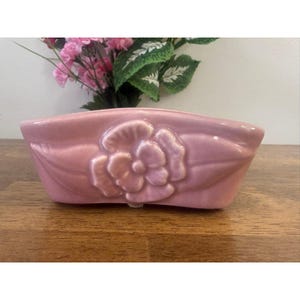 May include: A pink ceramic planter with a raised floral design. The rectangular planter has a curved shape and is decorated with a large, embossed flower in the centre. The planter is a soft pink colour.