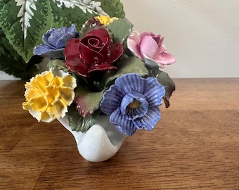 Puede incluir: Un arreglo floral de cerámica blanca con una variedad de flores coloridas. El arreglo incluye una rosa roja oscura, una rosa rosa, claveles amarillos y flores azules y moradas. Las flores están en un jarrón blanco curvado.