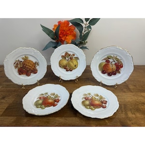 May include: Five white porcelain plates with scalloped edges and gold trim, each featuring a different fruit illustration. The fruits include a pineapple, apples, pears, and cherries. The plates are displayed on small gold stands.