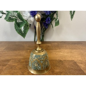 Może przedstawiać: Zabytkowy mosiężny dzwonek ze złotym uchwytem i dekoracyjną podstawą w turkusowe i złote wzory. Dzwonek jest eksponowany na drewnianej powierzchni, z bukietem kwiatów w tle. Dzwonek ma około 15 cm wysokości.