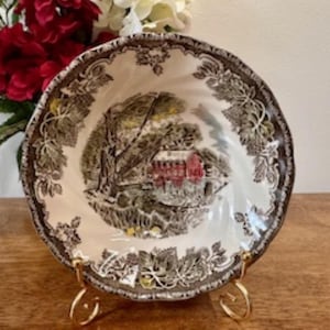 May include: A vintage ceramic bowl in off-white with a brown rim and a detailed landscape scene. The bowl is supported by a gold-coloured metal stand. The rim features a leaf pattern. The scene depicts a red building surrounded by trees.