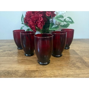 Peut inclure: Six verres à boire vintage teintés en rouge. Les verres ont une forme arrondie avec une base sombre et une teinte rouge plus claire vers le haut. Un bouquet de fleurs rouges et blanches est à l'arrière-plan.