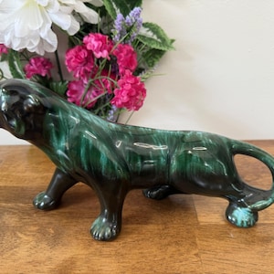 Peut inclure: Figurine de panthère en céramique avec glaçure verte et noire brillante. La figurine est en position de chasse, gueule ouverte, queue recourbée. Posée sur une surface en bois, avec des fleurs en arrière-plan.