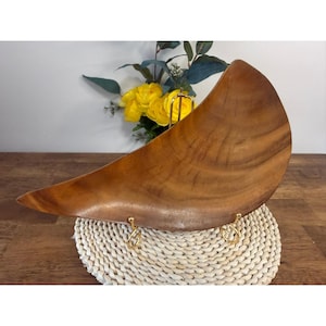 Puede incluir: Un cuenco de madera grande, en forma de media luna, con un acabado marrón intenso. El cuenco está sostenido por un soporte de metal dorado y descansa sobre un salvamanteles circular tejido. Se ven flores amarillas y vegetación en el fondo.