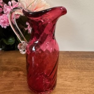 May include: A crimson glass jug with a clear handle and spout. The jug features a decorative swirl pattern and is likely for serving drinks. The jug is on a wooden surface.