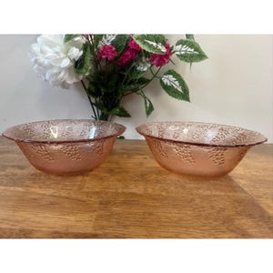 May include: Two pink glass bowls with a grape design. The bowls are oval-shaped with a slightly flared rim. The grape design is embossed on the exterior of the bowls. The bowls are sitting on a wooden surface.