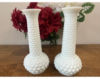 Puede incluir: Dos jarrones blancos de cristal hobnail, con base bulbosa y cuello estrecho. Los jarrones son de vidrio opaco y tienen una superficie texturizada. Flores rojas y blancas en el fondo. Los jarrones están sobre una superficie de madera.