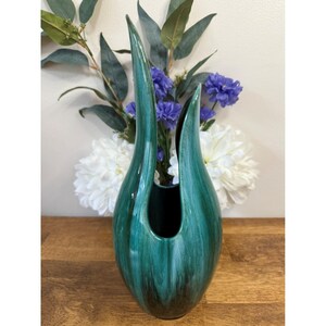 Peut inclure: Un vase en céramique de couleur teal, de forme haute et ouverte. Le vase a une finition brillante et une base marron foncé. Il est posé sur une surface en bois, avec des fleurs blanches et violettes en arrière-plan.