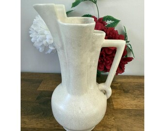 May include: Cream-colored vintage pitcher with a unique stepped design and a rounded base. The pitcher has a handle and a textured surface. Decorative flowers are in the background.