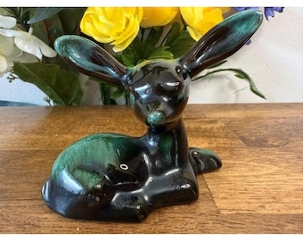 May include: A ceramic deer figurine in a resting pose. The deer has a glossy black finish with green accents on the ears, back, and nose. The figurine is displayed on a wooden surface with yellow flowers in the background.