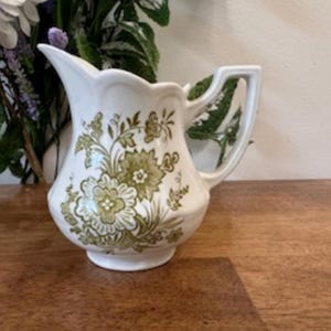 May include: A white ceramic jug featuring a green floral pattern. The jug has a decorative scalloped rim and a handle. The floral design includes stylized flowers and foliage. It is likely used for serving liquids, such as water or milk.