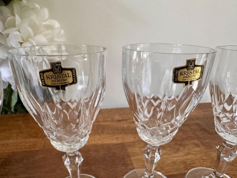 May include: Set of four clear glass wine glasses with a cut crystal design. Each glass has a gold label that reads "Kristal Zaječar".