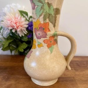May include: A beige ceramic pitcher with a floral design. The pitcher has a wide mouth and a handle. The flowers are painted in shades of orange, yellow, purple, and green.