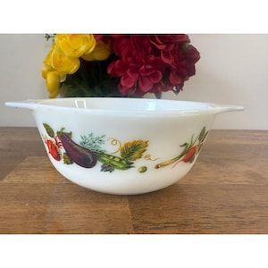 May include: White mixing bowl with a colourful vegetable design. The bowl features illustrations of a tomato, aubergine, peas, and other vegetables. The bowl has two handles and is set against a wooden surface.