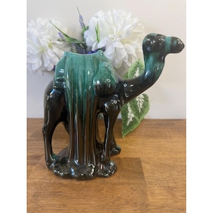 以下が含まれることがあります： 光沢のある黒とティールグリーンのセラミックラクダの置物。ラクダの背中には鞍のような開口部があり、おそらく花などを入れるため。置物は木製の表面に置かれています。