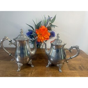 May include: Two silver teapots with decorative handles and lids. The teapots have a classic design with decorative feet. A bouquet of orange, blue, and purple flowers sits behind the teapots on a wooden surface.