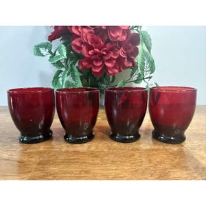 May include: Four red glass tumblers with a dark base are arranged on a wooden surface. The glasses have a rounded shape and are filled with a dark red colour. A bouquet of red flowers is in the background.