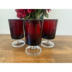 May include: Three matching drinking glasses with deep red bowls and clear glass stems and circular bases. The glasses feature a straight-sided, cylindrical bowl shape and are arranged in a cluster, showcasing the vibrant red glass color.