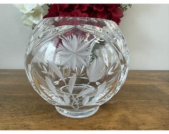 May include: A clear, round crystal vase with etched floral and leaf designs. The vase has a wide opening and sits on a short, rounded base. The vase is filled with red and white flowers, creating a decorative display.