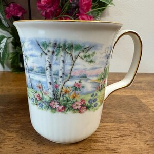 May include: White ceramic mug with a landscape scene of birch trees, a lake, and flowers. The rim and handle are trimmed with gold. A decorative item for the home.