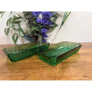 May include: Two rectangular, emerald green glass trays with textured sides and bottoms. The trays are displayed on a wooden surface, with a backdrop of purple flowers and green foliage.
