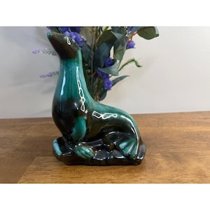 Peut inclure: Une figurine de phoque en céramique avec une finition brillante aux nuances mélangées de brun foncé et de vert sarcelle vif. Le phoque est représenté dans une posture assise et droite, la tête légèrement tournée, reposant sur une base sombre.