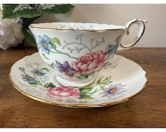 Può includere: Un set di tazza e piattino bianchi con un design floreale. La tazza e il piattino presentano fiori rosa, blu e viola con bordi dorati. La tazza ha un manico curvo e un bordo smerlato.