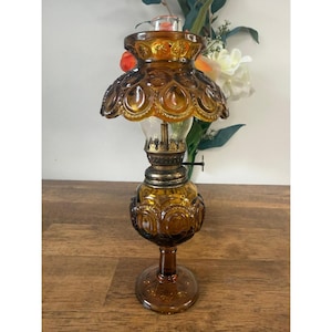 May include: Amber glass oil lamp with a decorative, scalloped shade and a round base. The lamp features a detailed, embossed design and a clear glass chimney. The lamp is approximately 30 cm tall.