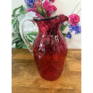 May include: A ruby red glass jug with a textured surface and a clear, twisted glass handle. The jug has a flared spout and a dark rim. Decorative flowers are in the background.