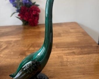 May include: A tall, glossy, emerald green ceramic swan figurine with a long, curved neck and a black base. The swan's head is black, and the body has detailed feather accents. The figurine is displayed on a wooden surface.