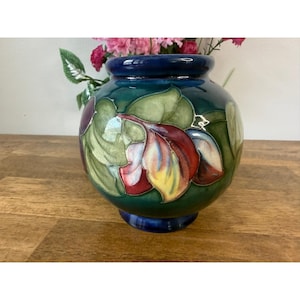 Puede incluir: Un jarrón de cerámica redondo con borde y base de color azul oscuro y acabado brillante. El cuerpo presenta un patrón en relieve de hojas grandes y superpuestas en tonos verde, rojo, amarillo y blanco, con un esmalte craquelado visible.