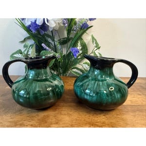 Peut inclure: Deux pichets en céramique verts et noirs assortis avec des anses. Les pichets ont une finition brillante et un dégradé vert foncé à noir. Ils sont posés sur une surface en bois avec des fleurs en arrière-plan.