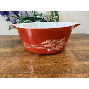 Könnte beinhalten: Eine rote Pyrex-Rührschüssel mit weißem Innenraum und Weizenmuster. Die Schüssel hat eine runde Form mit einem ausgestellten Rand und zwei Griffen. Das Weizendesign ist cremefarben.