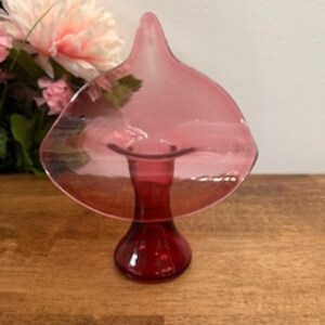 May include: A vintage, ruby-coloured glass centrepiece with a flared, petal-like top and a rounded base. The glass has a translucent quality, and the piece sits on a wooden surface. Decorative flowers are in the background.