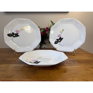 May include: A set of white octagonal plates and a bowl with a floral design. The plates and bowl have a black, pink, and yellow floral pattern. The plates are on gold stands, and the bowl is on a wooden surface.