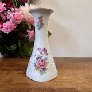 May include: White porcelain vase with a flared top and floral design. The vase features a delicate embossed pattern and is adorned with clusters of pink, purple, and blue flowers. The vase is set on a wooden surface.