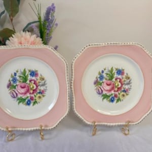 May include: Two square pink and white floral dinner plates with gold trim. The plates have a pink border and a white center with a floral design.
