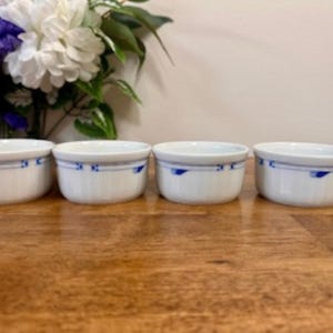 Puede incluir: Cuatro ramequines de cerámica blanca con un diseño floral azul alrededor del borde. Los ramequines están dispuestos en fila sobre una superficie de madera. Un ramo de flores blancas y moradas está en el fondo.