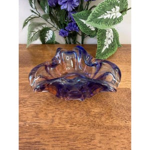 May include: A vintage glass ashtray with a unique, organic shape. The ashtray features a blend of purple and amber hues, creating a striking visual effect. The edges are gently curved, adding to its elegant design. A decorative piece for a home.