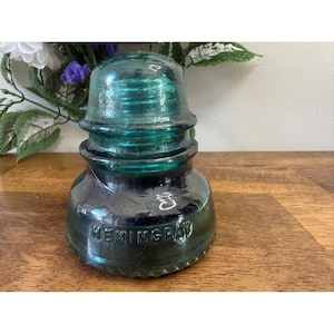 Könnte beinhalten: Ein Vintage-Glasisolator von Hemingray in Teal-Grün. Das Glas hat eine abgerundete Oberseite und einen breiteren Sockel mit dem geprägten Wort "HEMINGRAY". Er steht auf einer Holzoberfläche. Misst etwa 10 cm hoch.