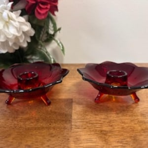 May include: Two ruby red glass candle holders. Each candle holder has a flower-shaped design with a scalloped edge and three small feet. The candle holders are on a wooden surface.