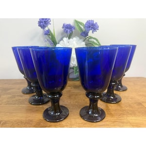 Puede incluir: Juego de seis copas de vino azul cobalto. Cada copa tiene un cuenco redondeado y un tallo y base de color marrón oscuro. Las copas están dispuestas sobre una superficie de madera, con un arreglo floral en el fondo.