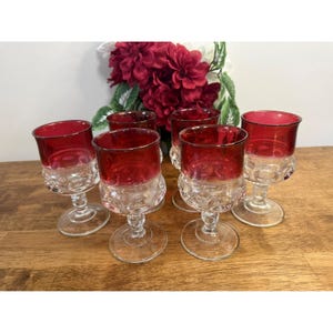 Puede incluir: Juego de seis copas de vino vintage. Cada copa presenta una base transparente estampada y un cuenco rojo vibrante con borde plateado. Las copas están dispuestas sobre una superficie de madera, con un arreglo floral al fondo.