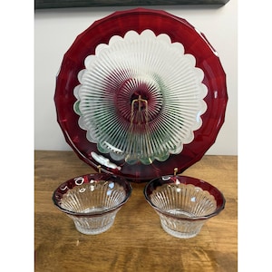 May include: A tiered serving set featuring a large red glass plate with a scalloped edge, a clear glass plate, and two small bowls. The set has a gold-colored metal stand. The bowls and plates have a red trim.