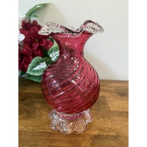 May include: A ruby red glass vase with a ruffled rim and a textured, swirled body. The vase sits on a clear, scalloped base. The background includes a bouquet of red flowers and green leaves, set against a wooden surface.