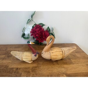 Pode incluir: Duas figuras decorativas de patos feitas de palha e madeira. Um pato tem pescoço vermelho e gravata borboleta xadrez. O outro pato tem pescoço de madeira. Um buquê de flores vermelhas e brancas está ao fundo.
