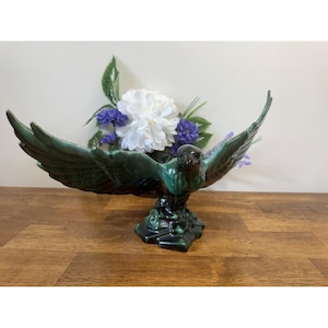 Könnte beinhalten: Keramische Adler-Skulptur mit ausgebreiteten Flügeln, glasiert in Grün- und Schwarztönen. Der Adler sitzt auf einem dekorativen Sockel und hält einen Strauß weißer und violetter Blumen. Ein dekoratives Stück für einen Tisch oder ein Regal.