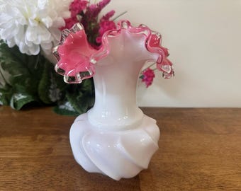 May include: A white glass vase with a ruffled pink rim. The vase has a bulbous base with a swirling design. The rim is a vibrant pink with clear edges. The vase is likely used for displaying flowers.